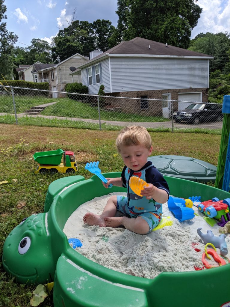 toddler sitting in green turtle sandbox and playing with sand toys